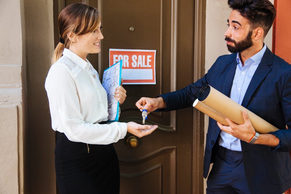 A woman receives house keys from a man in front of a door with a "For Sale" sign, both smiling.